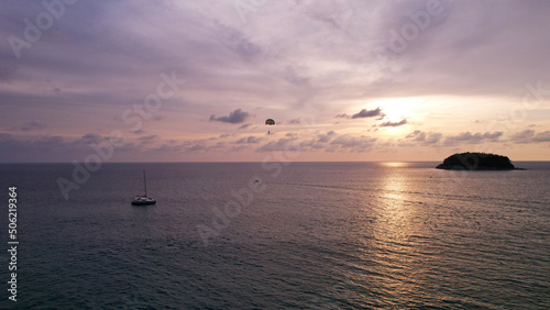 Parasailing at sunset with a view of the island. Parachute flight in the midst of a colorful sunset. Small waves, people relax on the beach, palm trees grow, there are hotels. Boats float. Asia Phuket © SergeyPanikhin