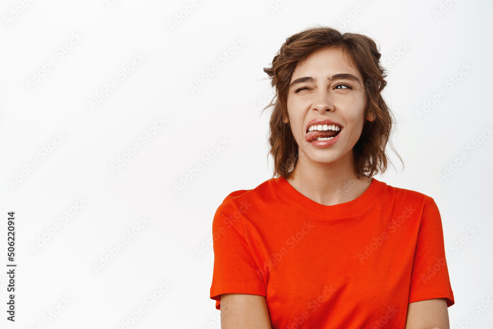 Portrait of attractive modern woman in red t-shirt, showing tongue, winning and smiling, looking at logo, advertisement, standing over white background Portrait of attractive modern woman in red t-shirt, showing tongue, winning and smiling, looking at logo, advertisement, standing over white background