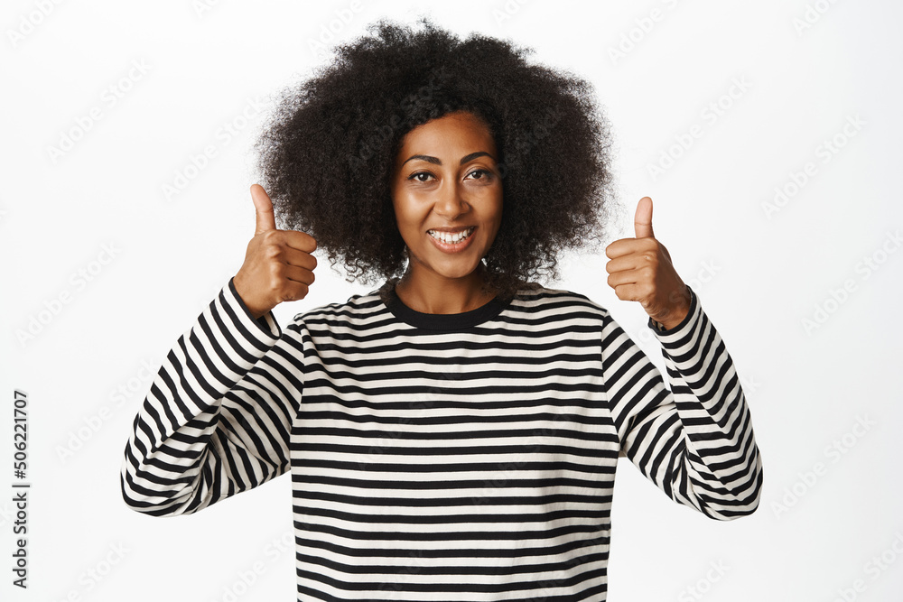 Portrait of smiling Black woman shows thumbs up in approval, like and agree, praise and compliment smth good, say yes, standing over white background Portrait of smiling Black woman shows thumbs up in approval, like and agree, praise and compliment smth good, say yes, standing over white background