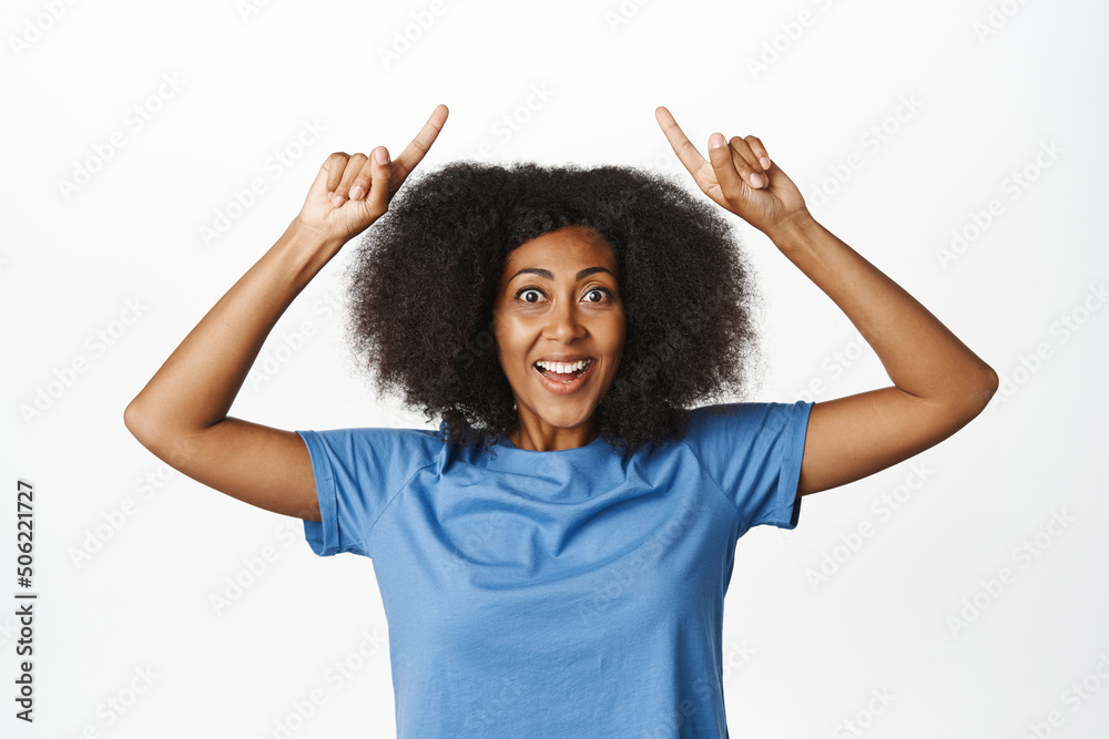 Enthusiastic african woman pointing fingers up, showing advertisement, information on top, making announcement with happy, excited smiling face, white background Enthusiastic african woman pointing fingers up, showing advertisement, information on top, making announcement with happy, excited smiling face, white background