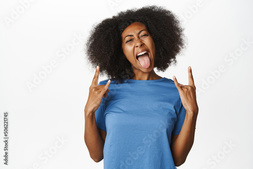 Enthusiastic Black female model, showing rock on, heavy metal party gesture, having fun, enjoying smth awesome, standing in blue t-shirt over white background