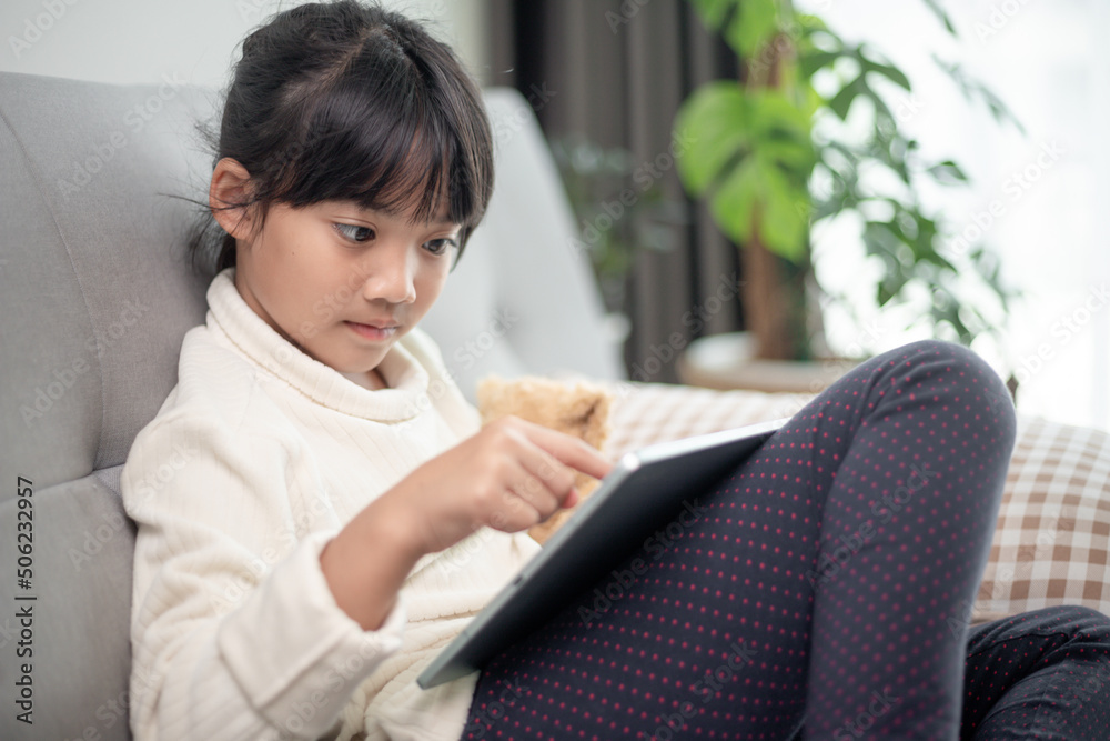 Little girl using tablet playing game on the internet, Kid sitting on sofa watching or talking with a friend online, Child relaxing in living room in the morning, Children with New Technology concept Little girl using tablet playing game on the internet, Kid sitting on sofa watching or talking with a friend online, Child relaxing in living room in the morning, Children with New Technology concept