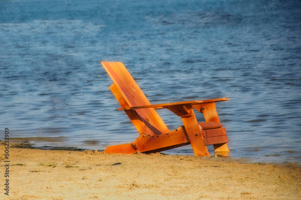 A new wooden Adirondack chair sits on the sandy beach at Cole Park in Upstate NY.  Beach chair by the water.