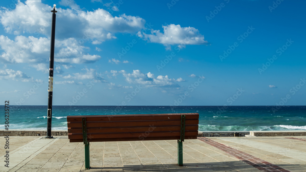 A wooden bench against the backdrop of blue cloudy sky and sea. 