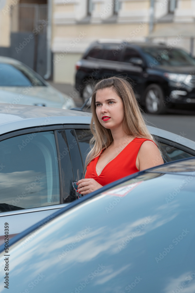 Young attractive girl wears red dress with deep neckline and red lipstick near cars. Vertical frame.