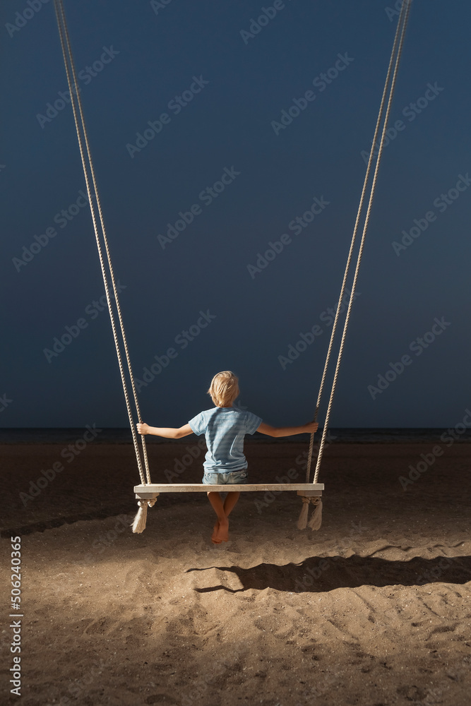 Portrait of boy on swing on the beach on dark sky background. Concept of childrens loneliness. Mystical back view portrait