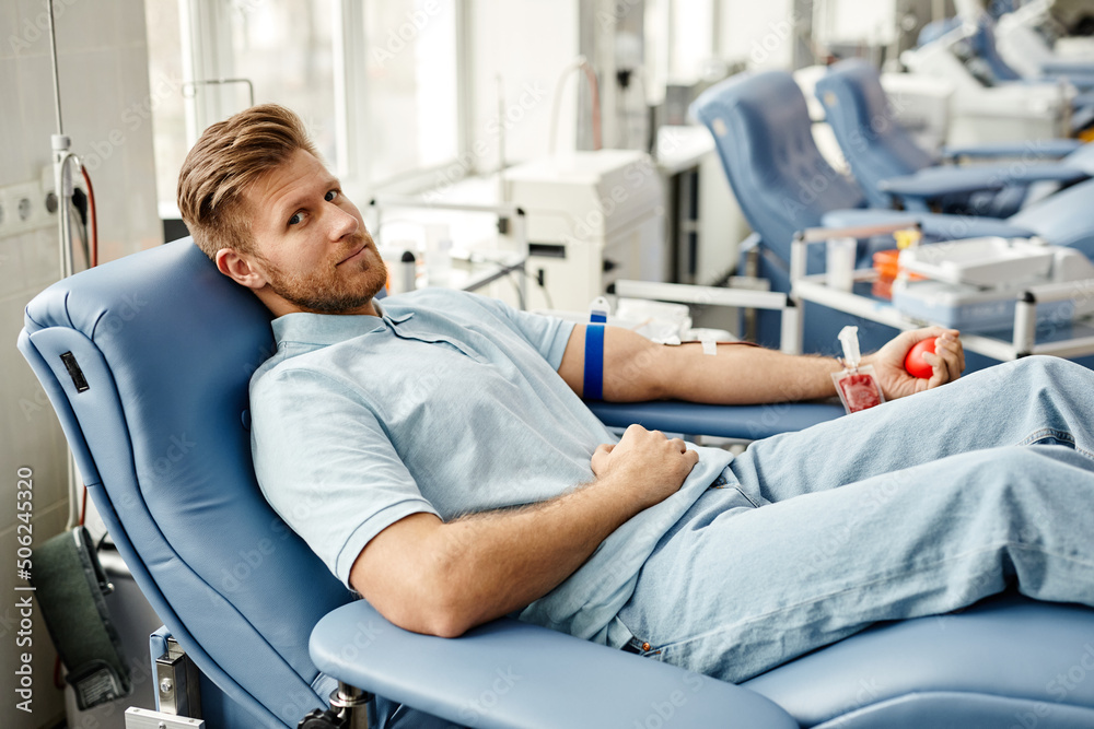 Full length portrait of young man donating blood while lying in comfortable chair at med center and looking at camera - obrazy, fototapety, plakaty 