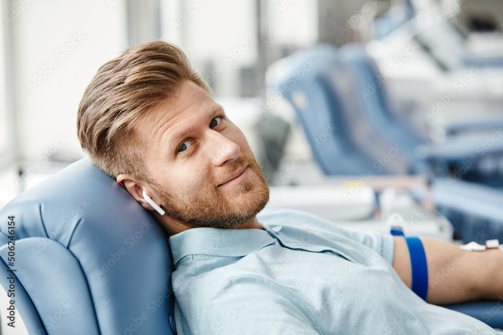 Portrait of smiling young man donating blood while lying in comfortable chair at med center and looking at camera - obrazy, fototapety, plakaty 