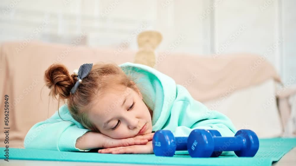 A primary school girl is bored with physical education. The schoolgirl looks sadly at the sports equipment and lies on the mat at home