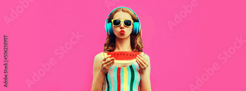 Summer portrait of stylish woman in headphones listening to music blowing her lips sends kiss with fresh juicy slice of watermelon on pink background, blank copy space for advertising text