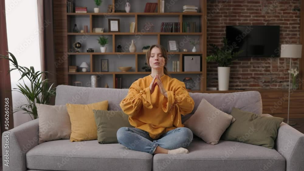 Beautiful woman meditating sitting on sofa in lotus pose with clised eyes in living room. Young girl doing yoga exercise at home for relaxing, stress relief, harmony and balance. Beautiful woman meditating sitting on sofa in lotus pose with clised eyes in living room. Young girl doing yoga exercise at home for relaxing, stress relief, harmony and balance.