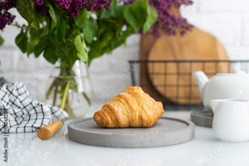 Fresh croissant on a grey plate on a table with a white teapot and lilacs