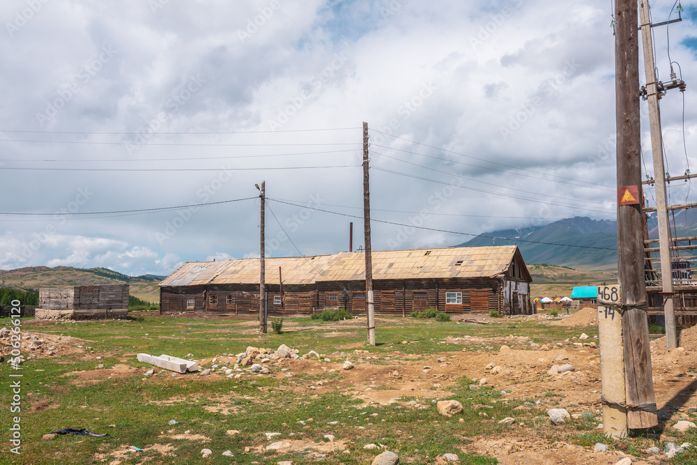 Countryside landscape with large long wooden barn with sloping roof among poles with wires in sunlight under cloudy sky at changeable weather in mountains. Sunlit old wide wood house under low clouds.