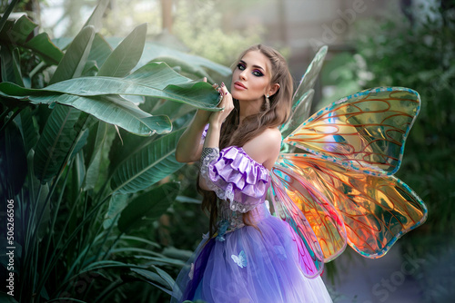A beautiful woman is a little cute fairy with butterfly wings in a green exotic forest. Fantasy shooting of fairy-tale characters. Forest nymph, pixie girl in purple dress