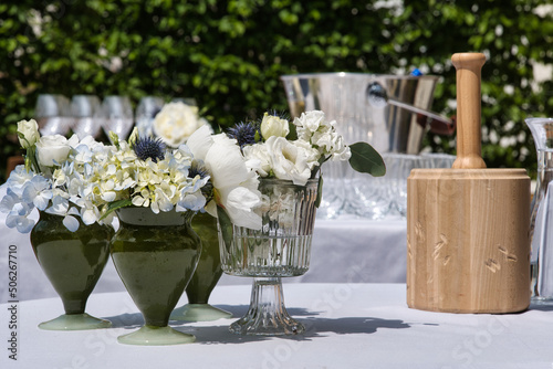 Flowers in vases on a table with a wooden hammer, which is used to open a beer barrel, next to it, and a wine cooler and glasses in the background. Wedding party setting. 