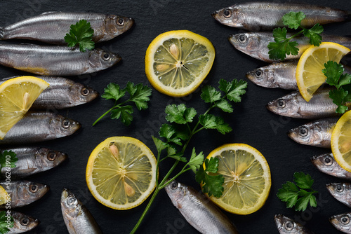 Small fish lie on a dark slate dish with sliced lemon and fresh parsley.