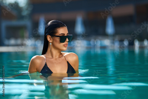 Fascinating brunette woman in sunglasses enjoying swimming in turquoise water of pool. Portrait view of gorgeous female looking away, while relaxing in pool of luxury hotel. Concept of wellness.