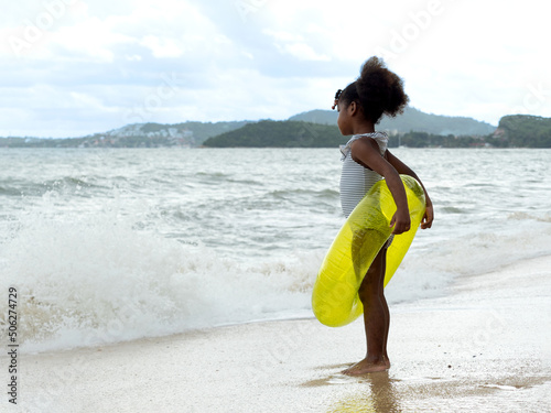 Little African girl wearing stripy swiming suit holding neon transparent swim ring, looking at the wave on the beach. Summer holiday concept.