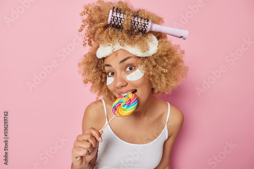 Lovely curly haired young woman bites multicolored lollipop has sweet tooth applies beauty patches to remove puffiness wears sleepmask and t shirt isolated over pink background. Daily routines