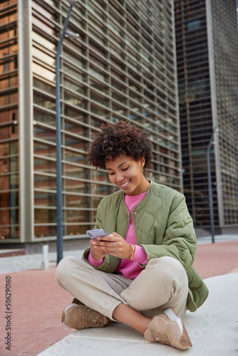 Cheerful young curly haired woman uses cellphone gadget for checking received email message browses website in social media wears jacket trousers and sneakers sits crossed legs in urban setting