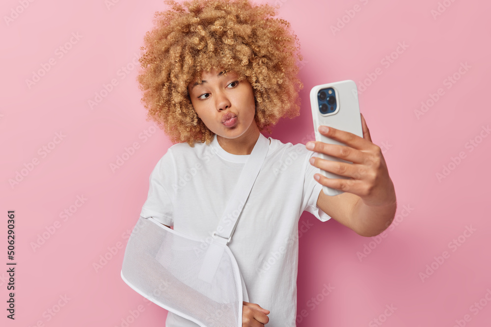 Injured woman with curly hair suffers from pain wears sling on arm keeps lips folded takes selfie via smartphone keeps lips folded dressed in casual white t shirt isolated over pink background Injured woman with curly hair suffers from pain wears sling on arm keeps lips folded takes selfie via smartphone keeps lips folded dressed in casual white t shirt isolated over pink background