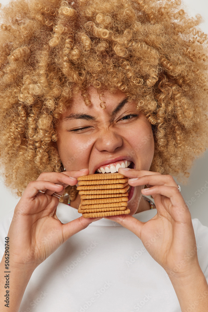 Vertical shot of curly haired beautiful woman bites pastries with appetite winks eye keeps mouth widely opened addiction to sugar and sweet food wears casual t shirt. Unhealthy eating concept Vertical shot of curly haired beautiful woman bites pastries with appetite winks eye keeps mouth widely opened addiction to sugar and sweet food wears casual t shirt. Unhealthy eating concept