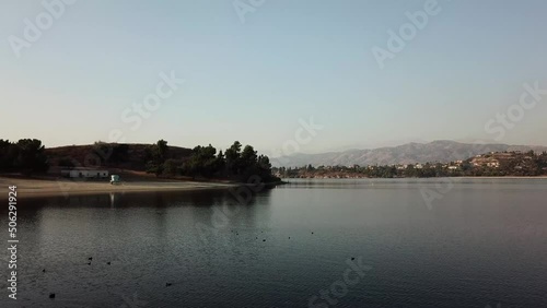 aerial footage of a vast blue rippling lake at sunset surrounded by lush green trees, grass and plants with majestic mountain ranges and blue sky at Puddingstone Lake in San Dimas California USA photo