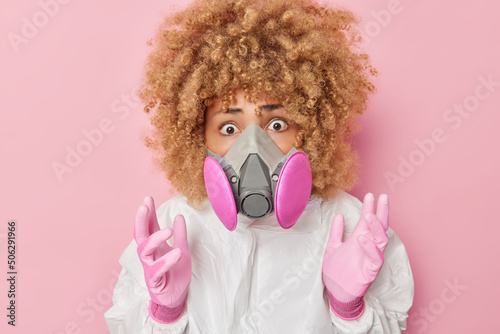Virus research. Worried curly haired woman keeps hands raised wears respirator and chemical suit reacts on shocking news isolated over pink background. Female scientist discovers coronavirus. © wayhome.studio 