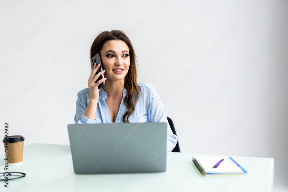 Good business talk. Cheerful young woman in glasses talking on mobile phone and using laptop with smile while sitting at her working place Good business talk. Cheerful young woman in glasses talking on mobile phone and using laptop with smile while sitting at her working place