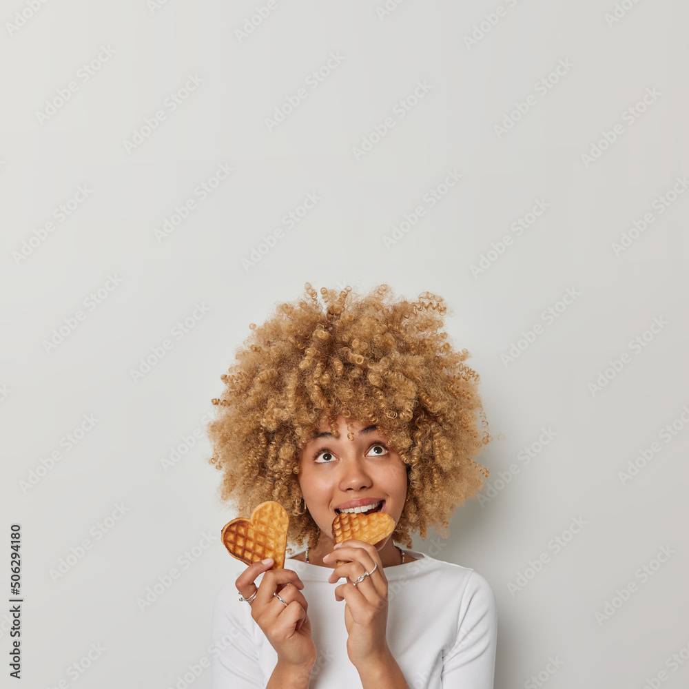 Vertical shot of lovely curly haired woman bites appetizing heart shaped waffles focused above enjoys eating delicious food dressed in casual jumper isolated over white background empty space Vertical shot of lovely curly haired woman bites appetizing heart shaped waffles focused above enjoys eating delicious food dressed in casual jumper isolated over white background empty space