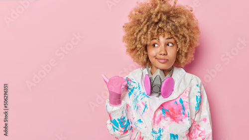 Curious young woman with curly hair wears safety suit and rubber gloves points thumb away on copy space tells you about biochemical hazard and virus protection isolated over pink background.