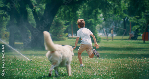 Cheerful little kid running from adorable pet at water sprinklers in summer park