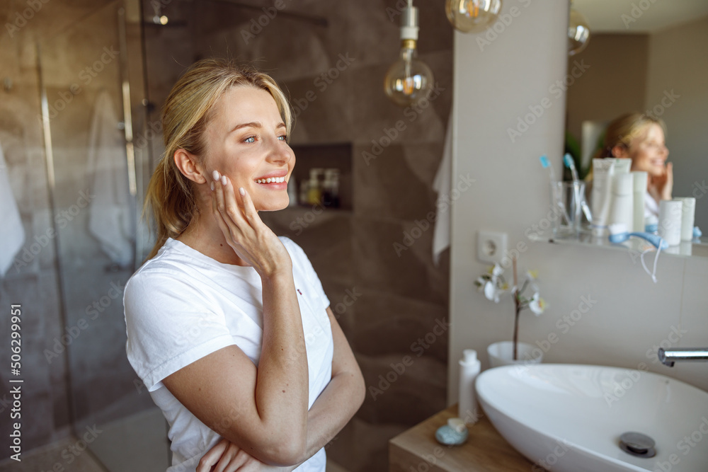 Dreamy young beautiful blonde woman smiling at mirror in bathroom. Self care concept. Dreamy young beautiful blonde woman smiling at mirror in bathroom. Self care concept.