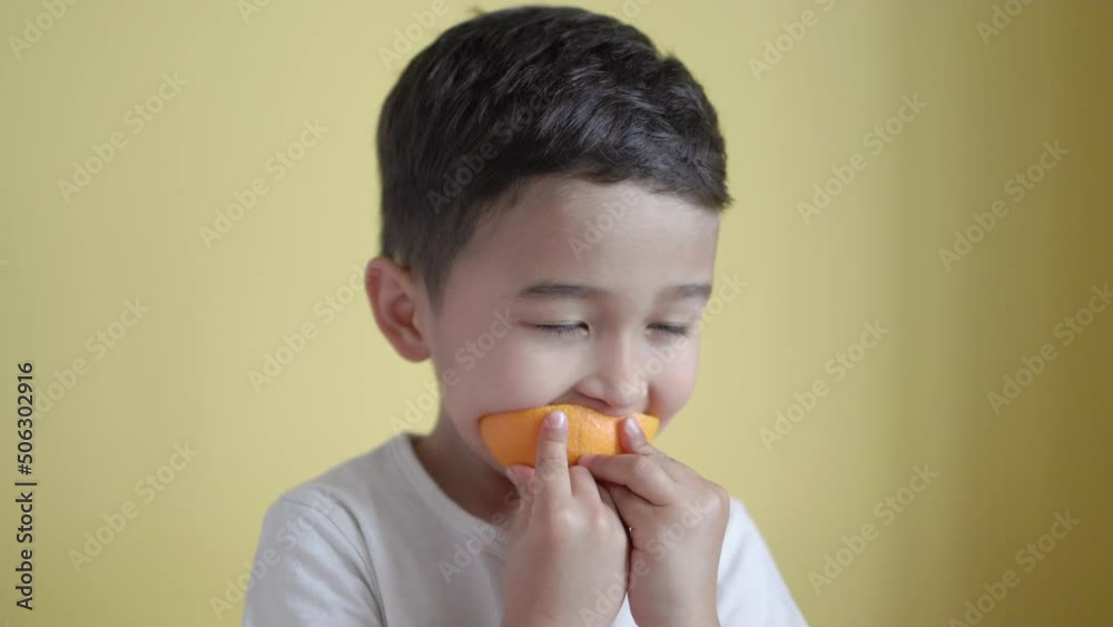 Cute asian boy eats a piece of orange on colour background Cute asian boy eats a piece of orange on colour background