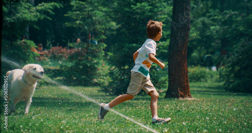 Active little boy running playing with dog on field with sprinklers on sunny day