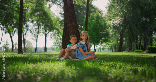 Smiling siblings play on grass under tree. Happy children sitting on lawn. © stockbusters
