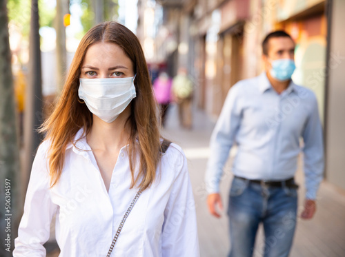 Focused young woman in medical mask walking to work along city street on spring day. New life reality during coronavirus pandemic.