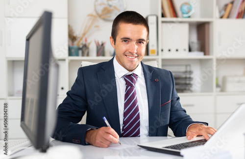 Portrait of smiling office employee sitting at desk, working with laptop and documents