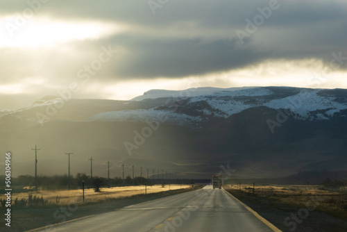 carretera asfaltada alumbrada por rayos de sol al atardecer saliendo de entre las nubes nimbostrato con cerros altos  nevados de fondo y un camión de carga a lo lejos  photo