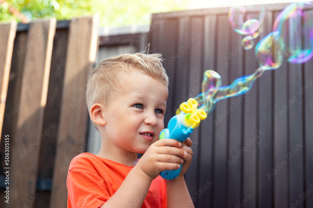 Close portrait of cute little bond kid boy enjoy having fun play blowing soap bubbles at home yard outdoors on bright warm summer day against house wall. Child healthy outside nature activities