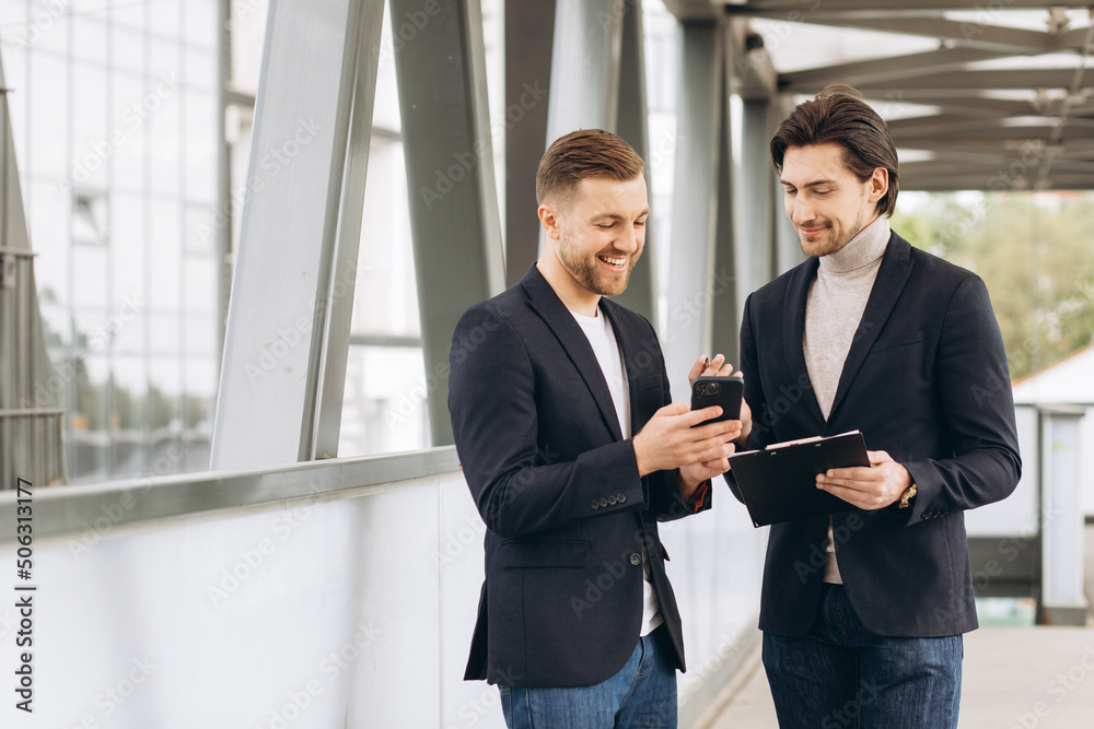 Two modern businessmen are discussing something and showing each other on a smartphone against the backdrop of urban offices and buildings Two modern businessmen are discussing something and showing each other on a smartphone against the backdrop of urban offices and buildings