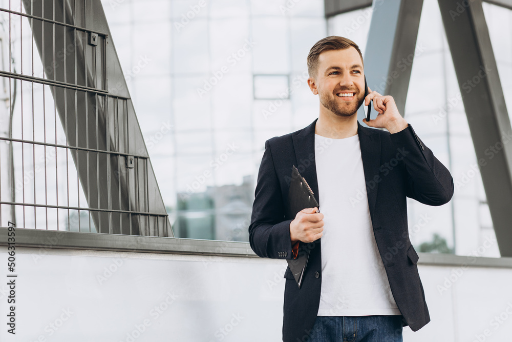 Portrait of happy modern male businessman in suit holding folder with documents and talking by phone against the background of urban buildings and offices. Portrait of happy modern male businessman in suit holding folder with documents and talking by phone against the background of urban buildings and offices.