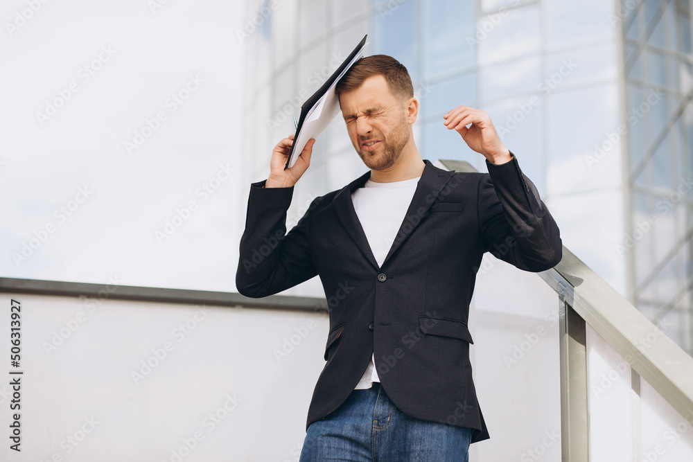 Portrait of a failed and stressed businessman hitting himself on the head with a folder of documents on the background of the office Portrait of a failed and stressed businessman hitting himself on the head with a folder of documents on the background of the office
