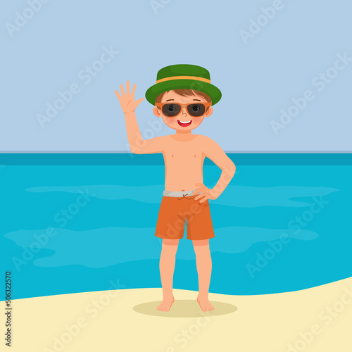 cute little boy in swimsuit with hat and sunglasses having fun on beach waving hand posing on summer vacation