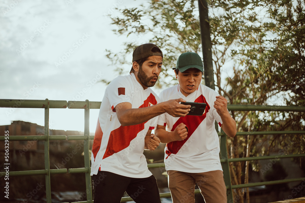 Dos fanáticos de Perú celebran un gol mirando su teléfono. Concepto de personas y deportes. - obrazy, fototapety, plakaty 