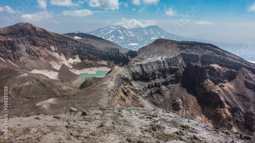 The top of an active volcano with a layered structure of rocky soil. There is a turquoise acid lake in the crater. Against the background of blue sky and clouds - a mountain range. Kamchatka. Gorely photo