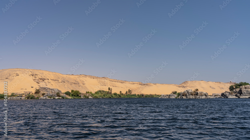 On the river bank there is a little green vegetation, picturesque boulders. High sand dunes against a clear sky. Ripples on calm blue water. Egypt. Nile