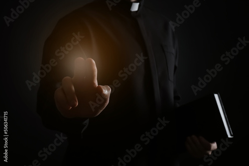 Priest with Bible making blessing gesture and holy light on dark background, closeup