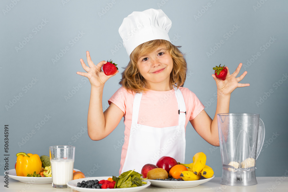 Child chef cook hold strawberries prepares food on isolated grey studio background. Kids cooking. Teen boy with apron and chef hat preparing a healthy vegetables meal in the kitchen. Child chef cook hold strawberries prepares food on isolated grey studio background. Kids cooking. Teen boy with apron and chef hat preparing a healthy vegetables meal in the kitchen.