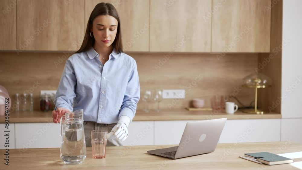 Young woman is working with laptop and pouring water at table in home kitchen spbas. 4k Beautiful European female with bionic hand looks at display and types text, pours fresh water into glass and Young woman is working with laptop and pouring water at table in home kitchen spbas. 4k Beautiful European female with bionic hand looks at display and types text, pours fresh water into glass and